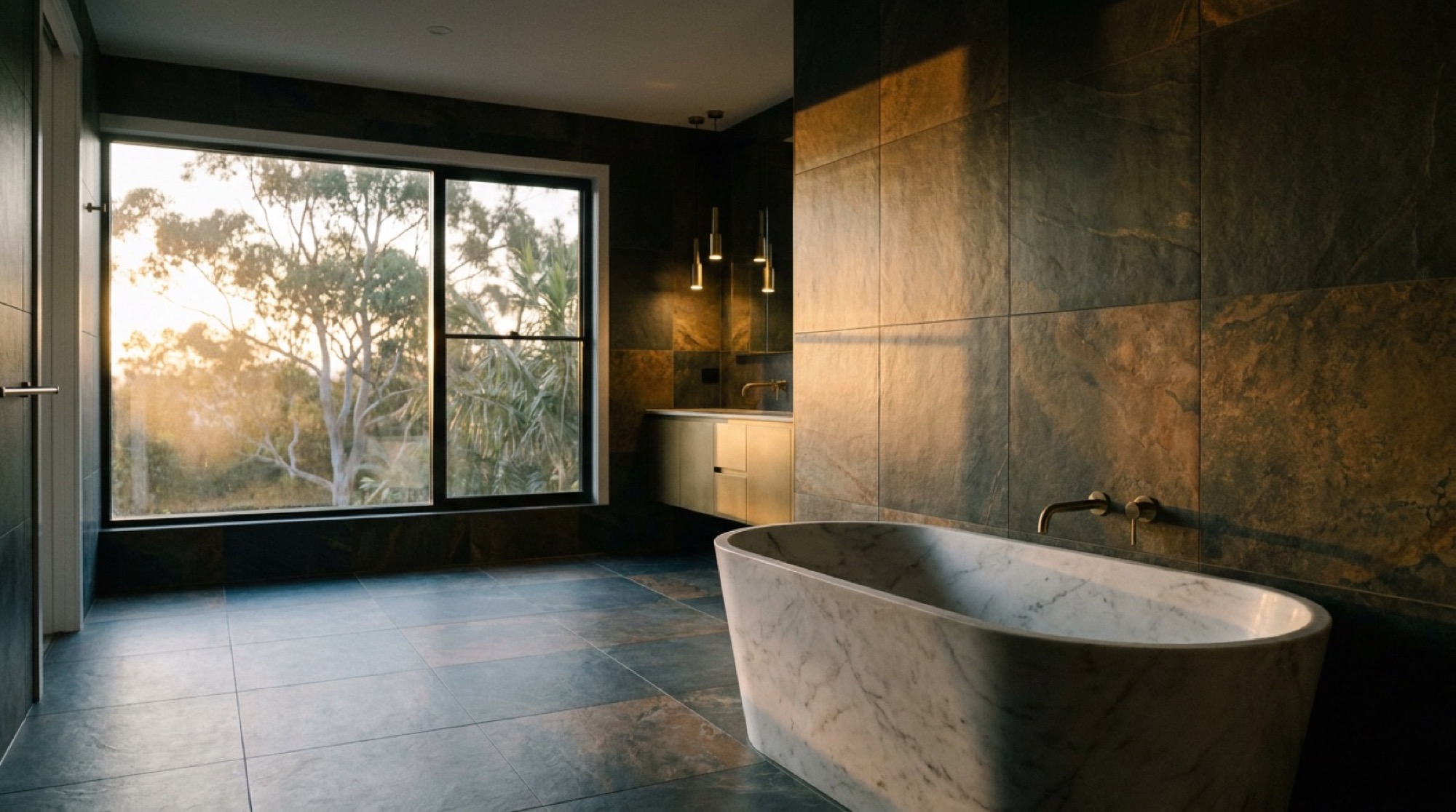 A dark stone tiled bathroom with a stone bath and a wide window looking into gum trees